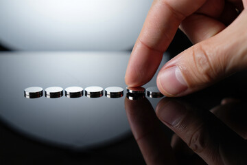 Hand Choosing Silver Button Battery from Line Displayed on Shiny Black Surface Reflecting White Light Concept Close up Shot with Selective Focus