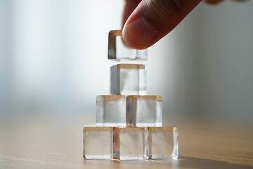 Hand Places Clear Acrylic Cubes in Pyramid Shape for Construction Building or Strength Concept 150 Characters
