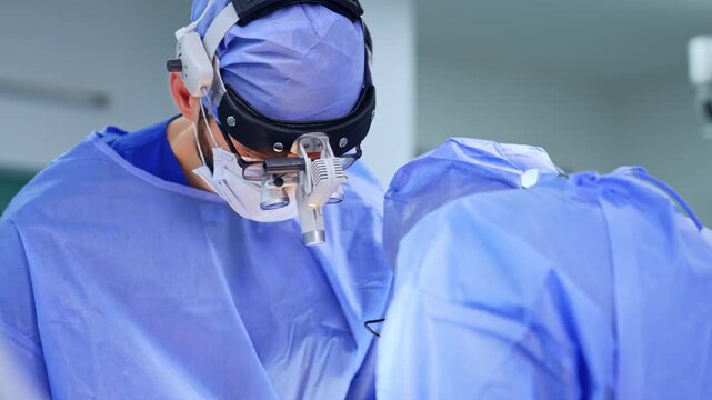 Unrecognized male surgeon wearing blue uniforms and surgical loupes leaned over the patient. Doctors work at neurosurgery.