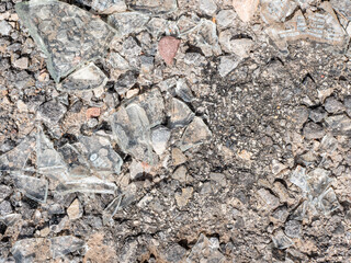 Shattered pieces of glass are mixed with gravel on a rough surface in a city setting. The debris suggests an area of neglect or a recent incident in broad daylight