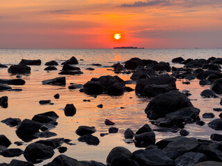 Serene sunset over a rocky shoreline, with the sun dipping into the horizon, casting warm hues across the sky and reflecting on the calm water. Tranquil summer evening at the beach.