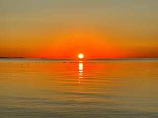 Beautiful sunset over the sea.  Bright sun touches the horizon, painting the expansive water and sky with vivid gold and amber tones. A calm, reflective scene captures the peaceful end of the day.