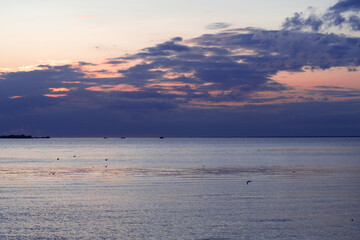 A serene coastal view at sunset, where dusky blue clouds contrast with pale pink light above the calm water. Small seabirds dot the peaceful horizon