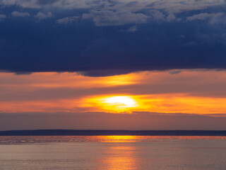 Scenic sunset over water displays vivid orange and yellow hues beneath heavy, dark clouds. The sun creates a glowing reflection on the calm surface. Dramatic cloudscape background.