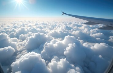 View from airplane window over dense cloud layer. Bright sun shines through fluffy white clouds against clear blue sky. Aircraft wing is visible.