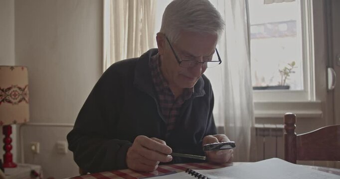 An elderly man with glasses intently reads a book or engages in a hobby at his home, using a magnifying glass to focus on the details. He appears calm and concentrated in a peaceful domestic setting.