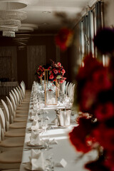 Elegant wedding table setup with a long white banquet table decorated with floral centerpieces in shades of red and pink. The arrangement features luxurious tableware, soft lighting, and a refined eve