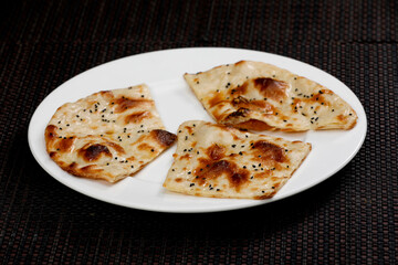 Three pieces of soft, fluffy Indian Naan or Kulcha flatbread, brushed with butter/ghee and sprinkled with black sesame seeds, arranged on a white plate against a dark woven background.