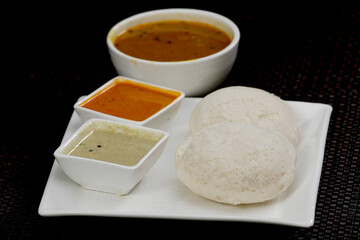Fluffy, soft South Indian Idli (steamed rice cakes) served with a traditional spread: rich Sambar (lentil stew) and two colorful chutneys (coconut and tomato) on a white platter.