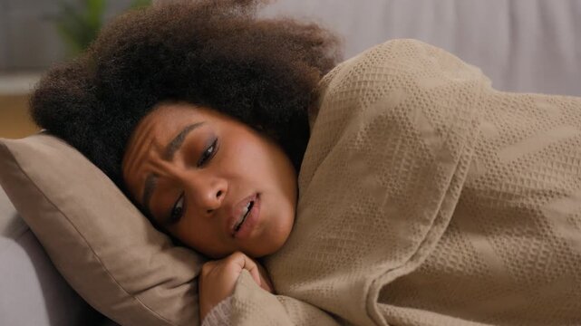 Frustrated ethnic African American female woman girl lying on comfortable cozy couch in living room relaxing on sofa feeling cold ill sick low temperature indoors covering blanket at home warming