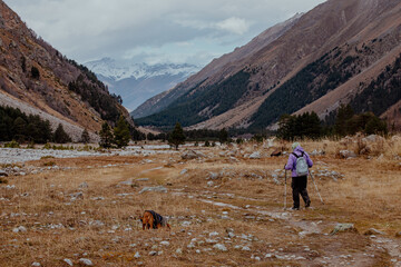 Kabardino-Balkaria, Russia-10.15.2025: Man walks his brown dog in a valley surrounded by mountains. The landscape features rocky terrain and sparse vegetation under a cloudy sky.