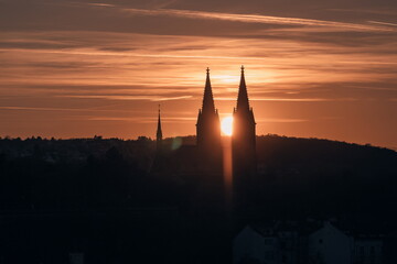 Obraz premium Dramatic sunset behind Prague’s Vyšehrad Basilica, the sun glowing perfectly between the twin spires and casting a warm orange silhouette over the historic skyline.