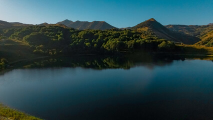 Aerial view of a serene lake surrounded by lush green hills and mountains in the Caucasus region. Clear blue sky reflects on the water's surface.