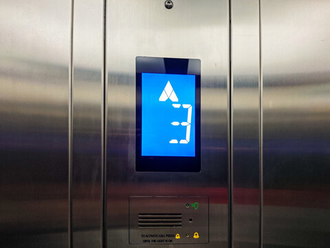 Close-up of a modern elevator panel displaying the number 3 and an upward arrow on a blue screen, indicating the current floor and direction. The brushed metal finish adds a sleek aesthetic.