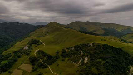 Aerial view of the Caucasus mountains. Rolling green hills and winding dirt paths are visible under a cloudy sky. The landscape is lush and expansive.