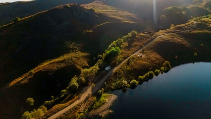 Aerial view of a serene landscape in the Caucasus region. Rolling hills, a winding road, and a calm lake are visible under bright sunlight.