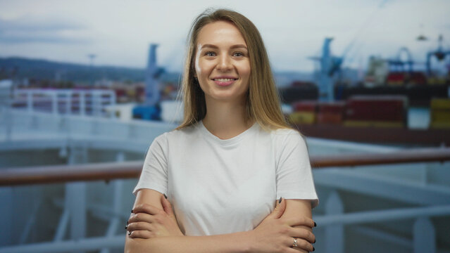 Woman stands confidently on a cruise boat in port, wearing a white shirt, with cargo containers in the background, exuding a sense of adventure and travel outdoors.