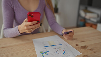 Woman holding smartphone and document in living room while analyzing data, surrounded by credit card and paperwork on wooden table, suggesting financial planning at home.