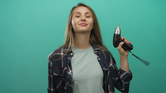 Young woman with long hair extending hand for handshake while holding drill against green background wall wearing casual plaid shirt and smiling confidently.