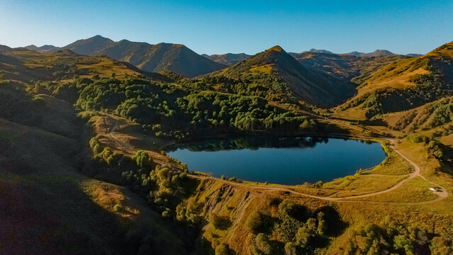 Aerial view of a serene lake surrounded by lush green hills and mountains in the Kavkaz region. Clear blue sky enhances the tranquil landscape.