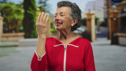 Senior woman with grey hair joyfully gesturing outdoors in an old town street, wearing a red dress, smiling, embodying happiness and nostalgia in a charming urban setting.