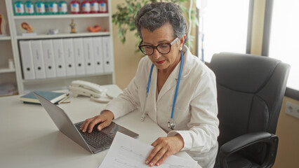 Senior woman doctor with grey hair working in office clinic on laptop wearing stethoscope surrounded by medical files and telephone in a professional healthcare environment.