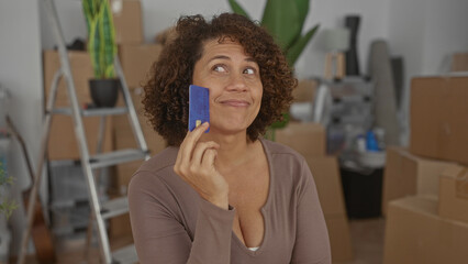 Woman holding creditcard to cheek while surrounded by moving boxes in building; thoughtful planning.