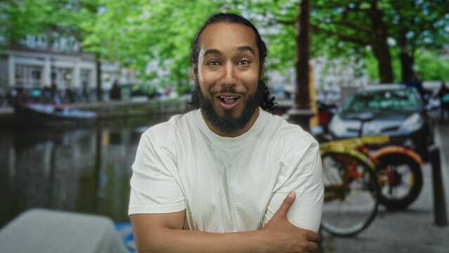 Man with clasped hands and open mouth on street by amsterdam canal with bicycle and water reflections; surprise travel.