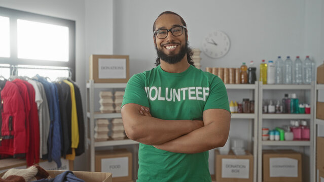 Man in green volunteer shirt standing with arms crossed by donation box in building; community service pride. - Powered by Adobe
