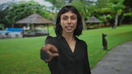 Woman raising bare hand with pointing finger and smiling face toward camera in natural forest;...