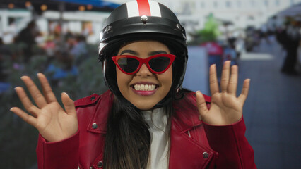 Young woman wearing helmet and red sunglasses smiles joyfully on an outdoor terrace, showcasing vibrant energy and urban style at a restaurant setting.