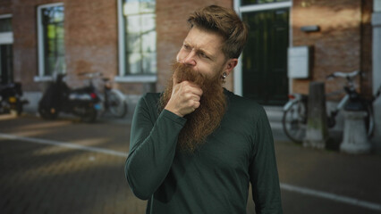 Naklejka premium Young bearded man grips his beard with hand while looking away in front of building; reflective curiosity.