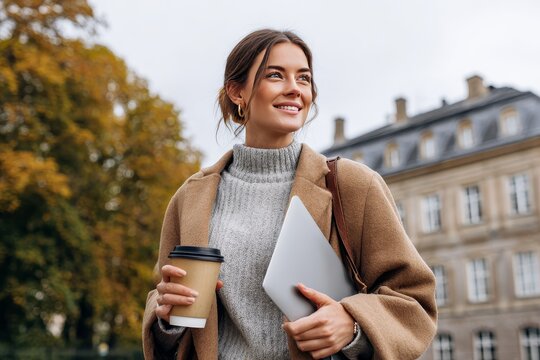 Smiling young female professor or business woman holding a laptop and coffee cup, leaving a historic university building on a cloudy autumn day