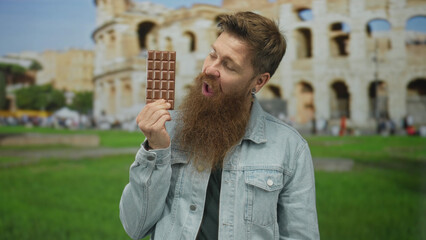 Man holds chocolate bar in front of ancient roman coliseum building while smiling broadly; joy...