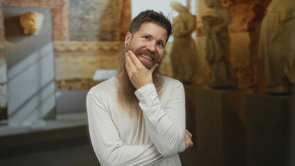 Obraz premium Bearded man with earrings touches his chin in a museum building among ancient marble statues under soft lighting; contemplation.