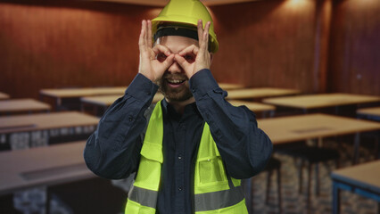 Obraz premium Man builder in a building wearing yellow hardhat and high visibility vest makes hand binoculars with fingers to eyes; safety training curiosity.