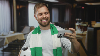 Man smiling and holding soccer ball and green scarf, holds phone for selfie with right hand in building; team pride joy.
