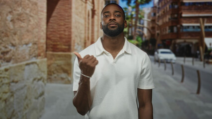 Man points thumb over shoulder on a city street, wearing white polo near brick building and parked...