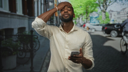 Man scrolling smartphone while walking along a sunlit city street lined with parked cars and trees;...