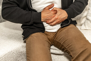 A young man in brown long pants sits on a white sofa in the living room, holding his stomach,