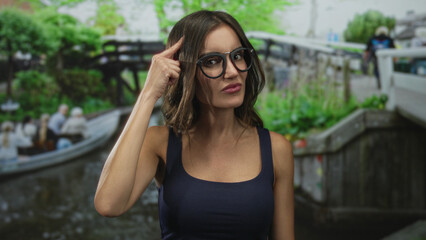 Young woman wearing glasses and a blue tank top raises finger near canal on street; curiosity discovery.