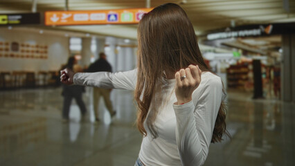 Young hispanic woman covering mouth with hand, arm outstretched, wearing white long sleeve shirt at airport terminal concourse; surprise travel delay.