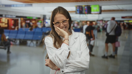 Woman scientist in white lab coat laughing with hand to chin at busy airport terminal indoors; joyful travel.