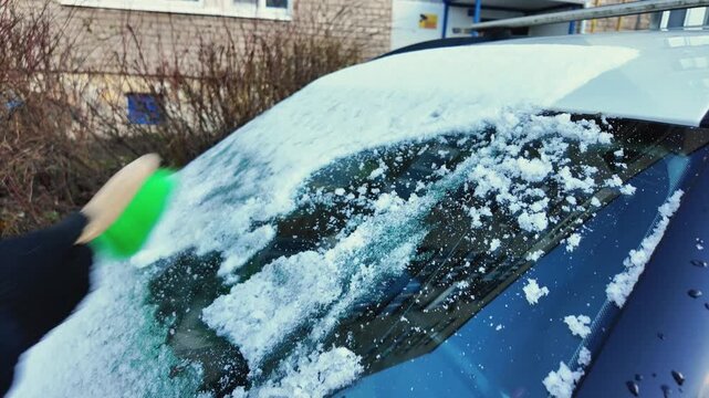 gloved hand scrapes icy windshield with green brush, compact car on residential street, brisk strokes fling snow chunks, cold blue light, safety-focused pre-drive routine, visible frost and melting