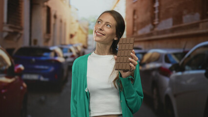 Woman holding a large chocolate bar with one hand on a narrow city street between parked cars,...
