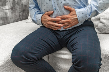 A man in elegant trousers and a long blue shirt sits on a white sofa, holding his aching stomach, hands on his stomach.