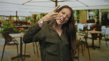Woman smiling and making rock horns gesture with raised hand at a restaurant terrace outdoors under umbrellas and tables; joy.