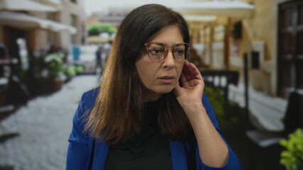 Woman wearing glasses touches her ear on a busy cobblestone street lined with outdoor cafe tables; concern.