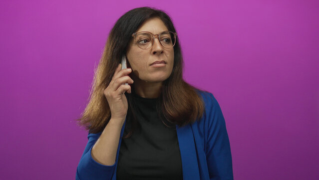Hispanic woman in blue blazer tilts head in side profile while holding smartphone to ear in studio; concentration.