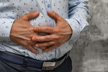 Close-up of a man's hands holding his aching stomach. A man wearing a blue long-sleeved shirt stands in a medical clinic